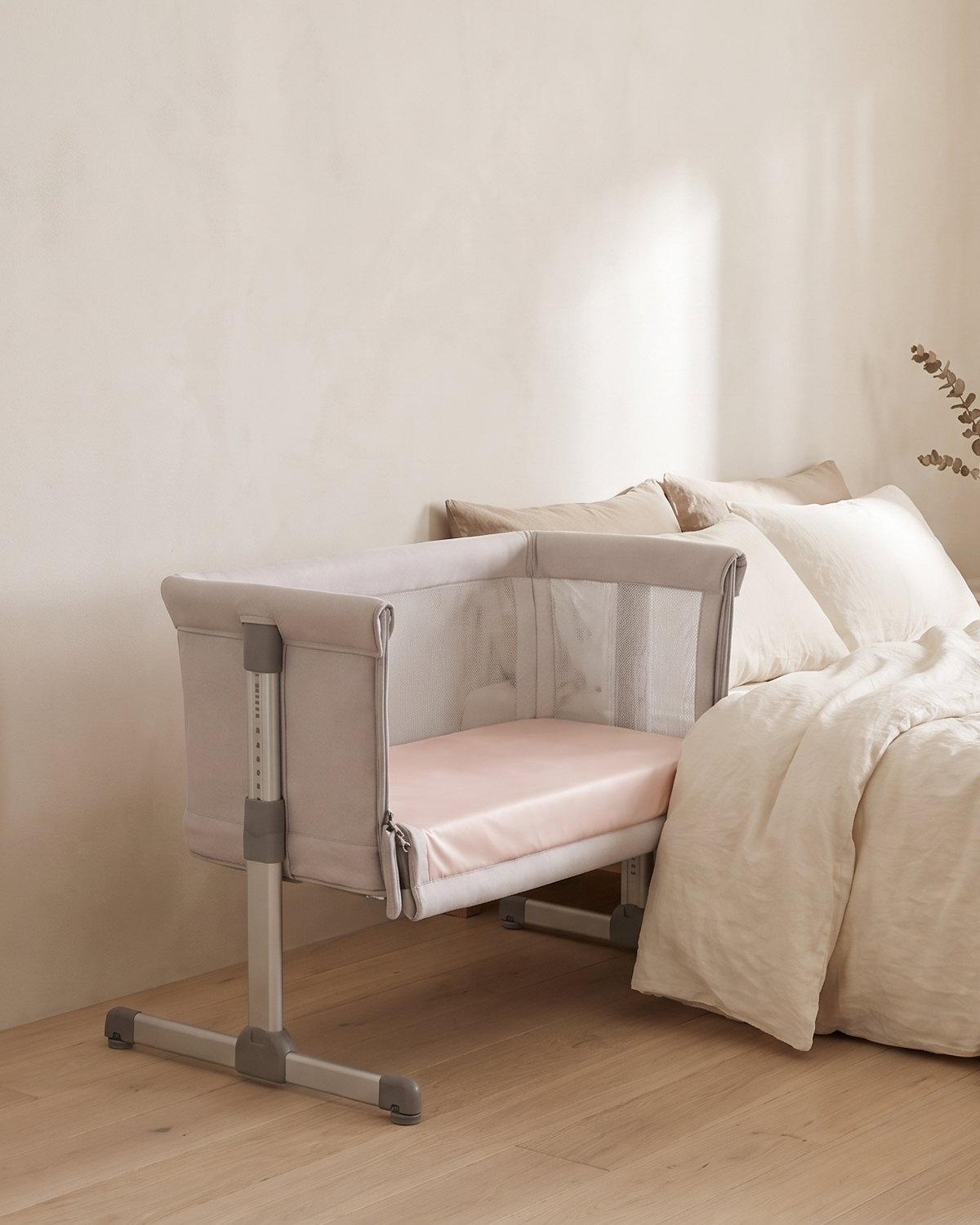 A bedside crib fitted with a Light Pink Sleepy Teddy silk sheet beside a bed